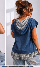 Woman wearing a blue sleeveless hoodie with a checkered bottom, standing against a white wall.