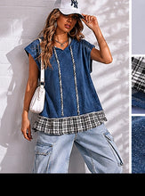 Woman wearing a blue layered top with plaid trim, white cap, and jeans against a light background.